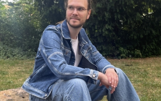 Pastor Peter Seeling mit hellblauer Jeans, weißen T-Shirt und blauer Jeansjacken sitzt draußen vor Büschen und Bäumen auf einem Stein und guckt nach links