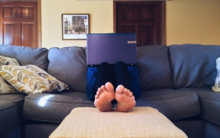 Mensch auf dem Sofa hinter dem Laptop im Vordergrund seine Füße